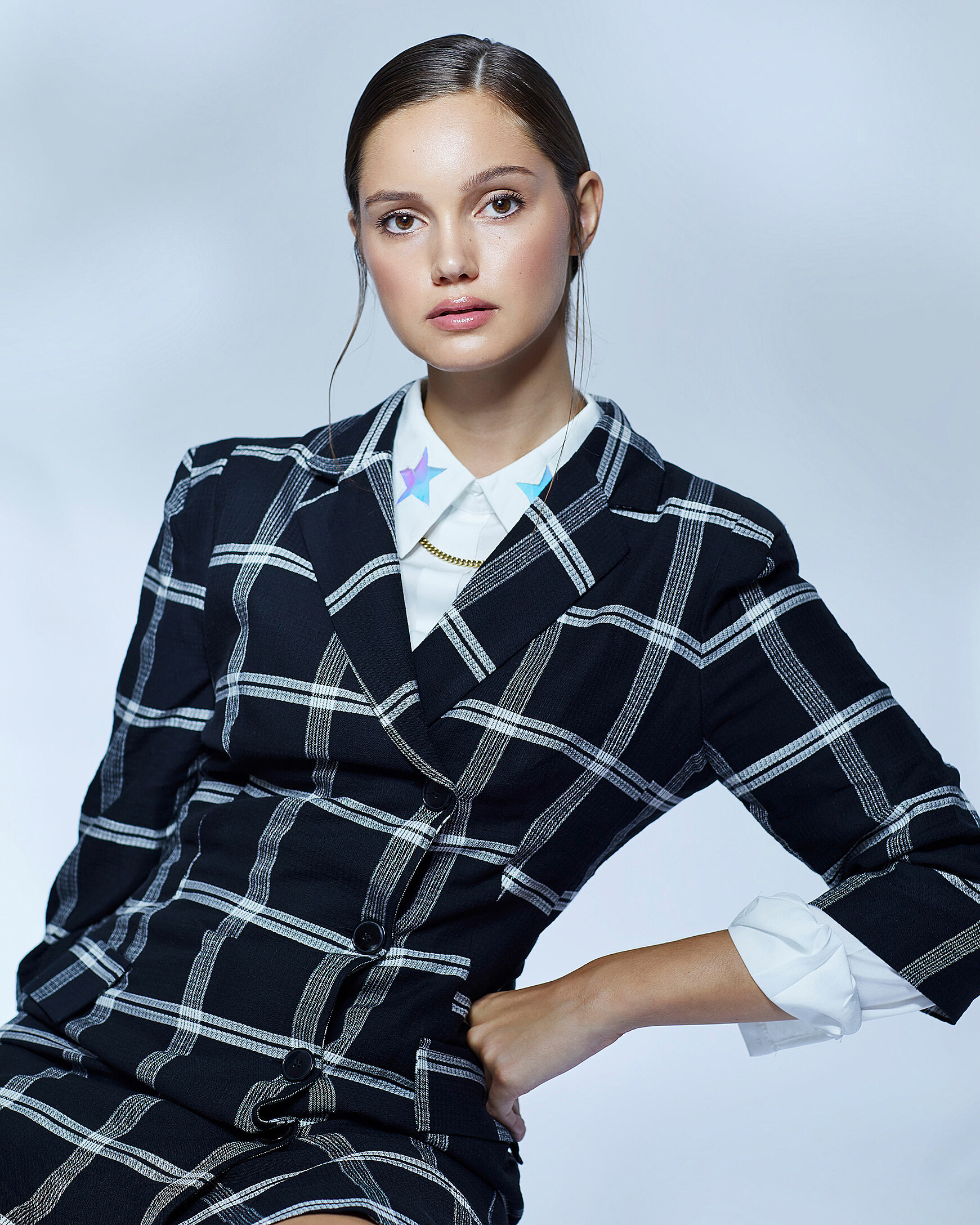 A female model who wears a checked blazer with a white blouse with blue stars on the chocker and sleek brown hair. She looks direct with a strong view into the camera.