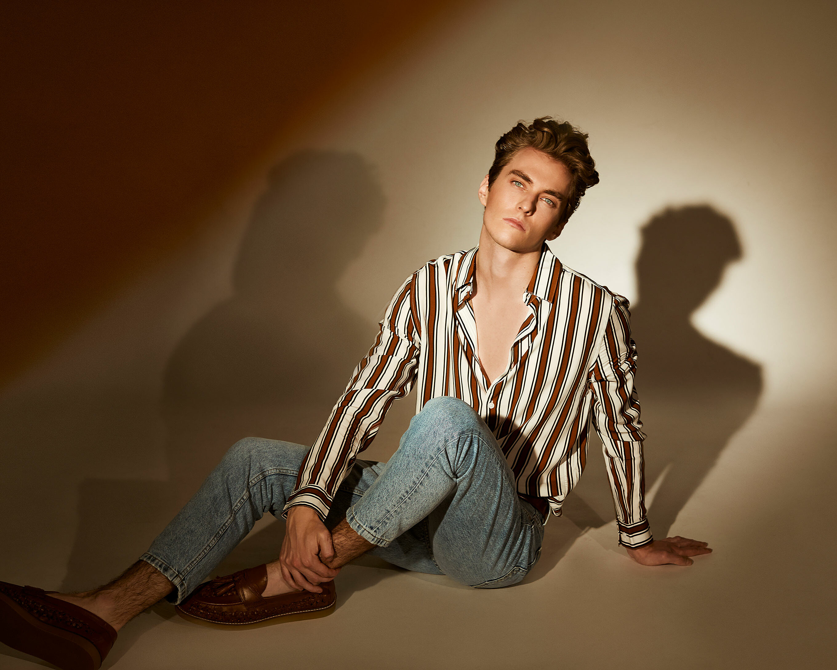A male model sits on the floor with a striped shirt and denim trousers