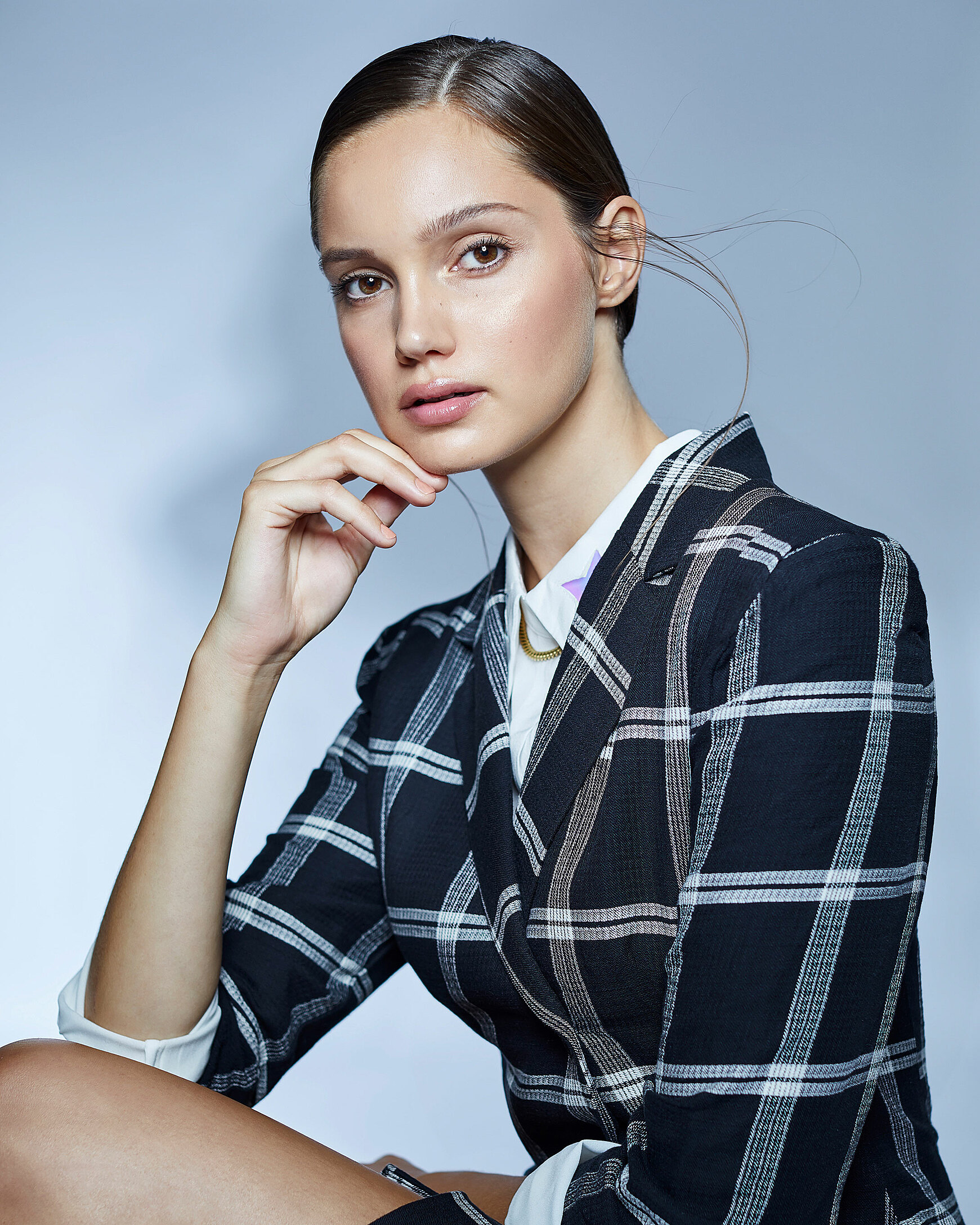 A female model who wears a checked blazer with a white blouse and sleek brown hair who sit in front of the camera