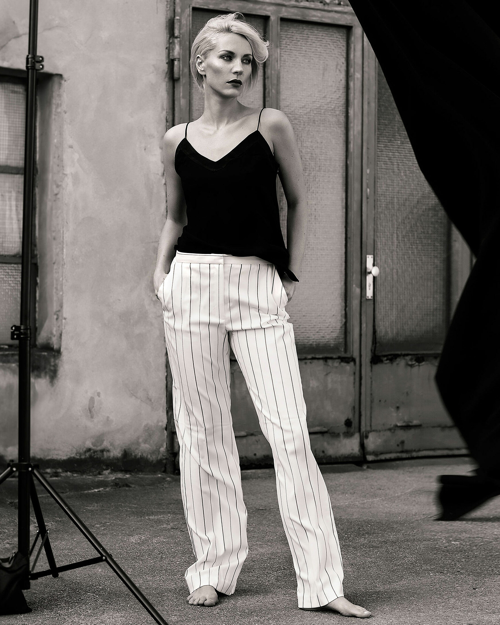 A black and white photo of a blonde woman in white long trousers with stripes and a black triangle top in front of a door in a backyard
