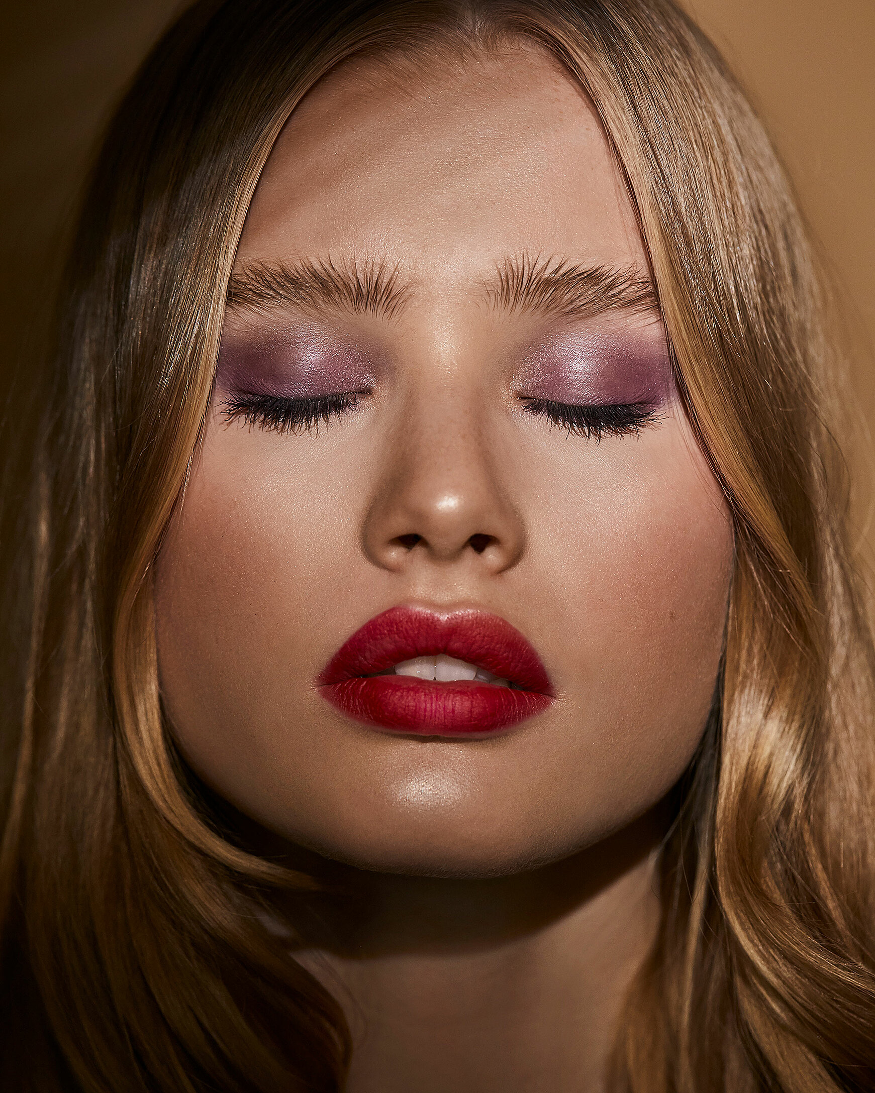 A close-up of a blonde female model closed her eyes. A colorful makeup with red lips and purple eye shadow