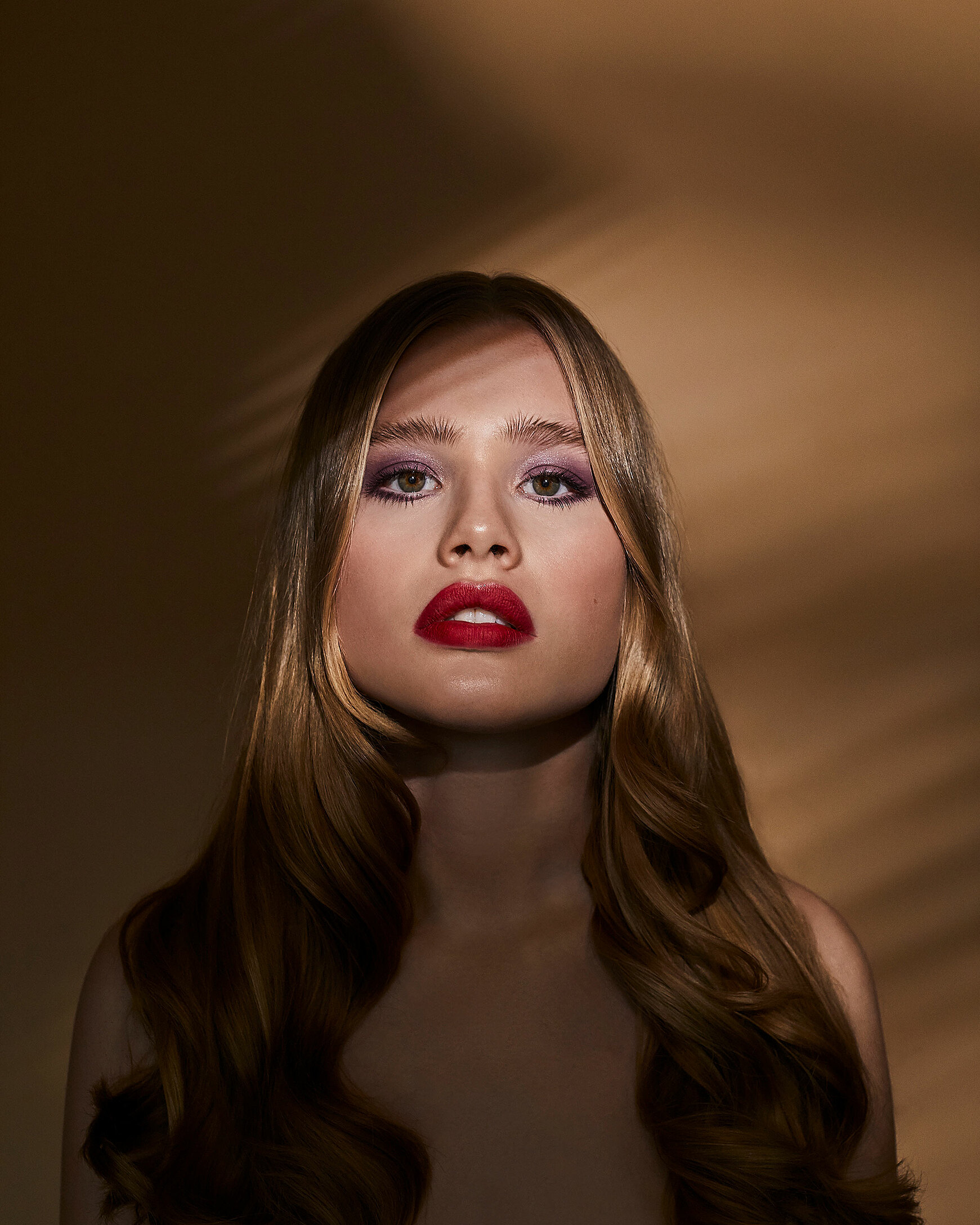 A female blonde model with red lips and purple eye shadow stand straight in front of the camera. The shadow is around her face