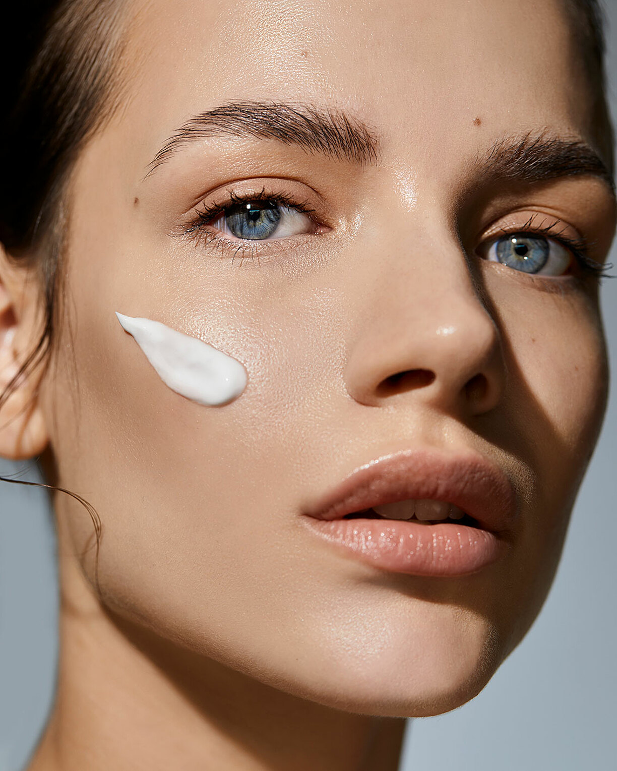 A beauty photography of a female model with dark hair. She has cream on her cheeck.