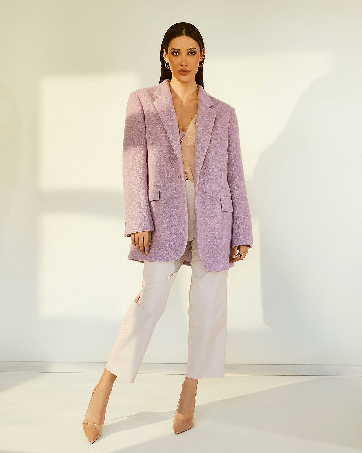 A female model stands in a sunlight from the window in rose trousers and a lilac coat