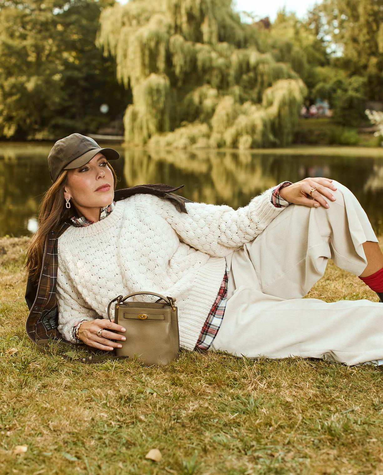 Eine Frau liegt auf einer Wiese vor einem See, Natur ist im Hintergrund zu sehen und sie posiert in beiger Mode mit einer Tasche.