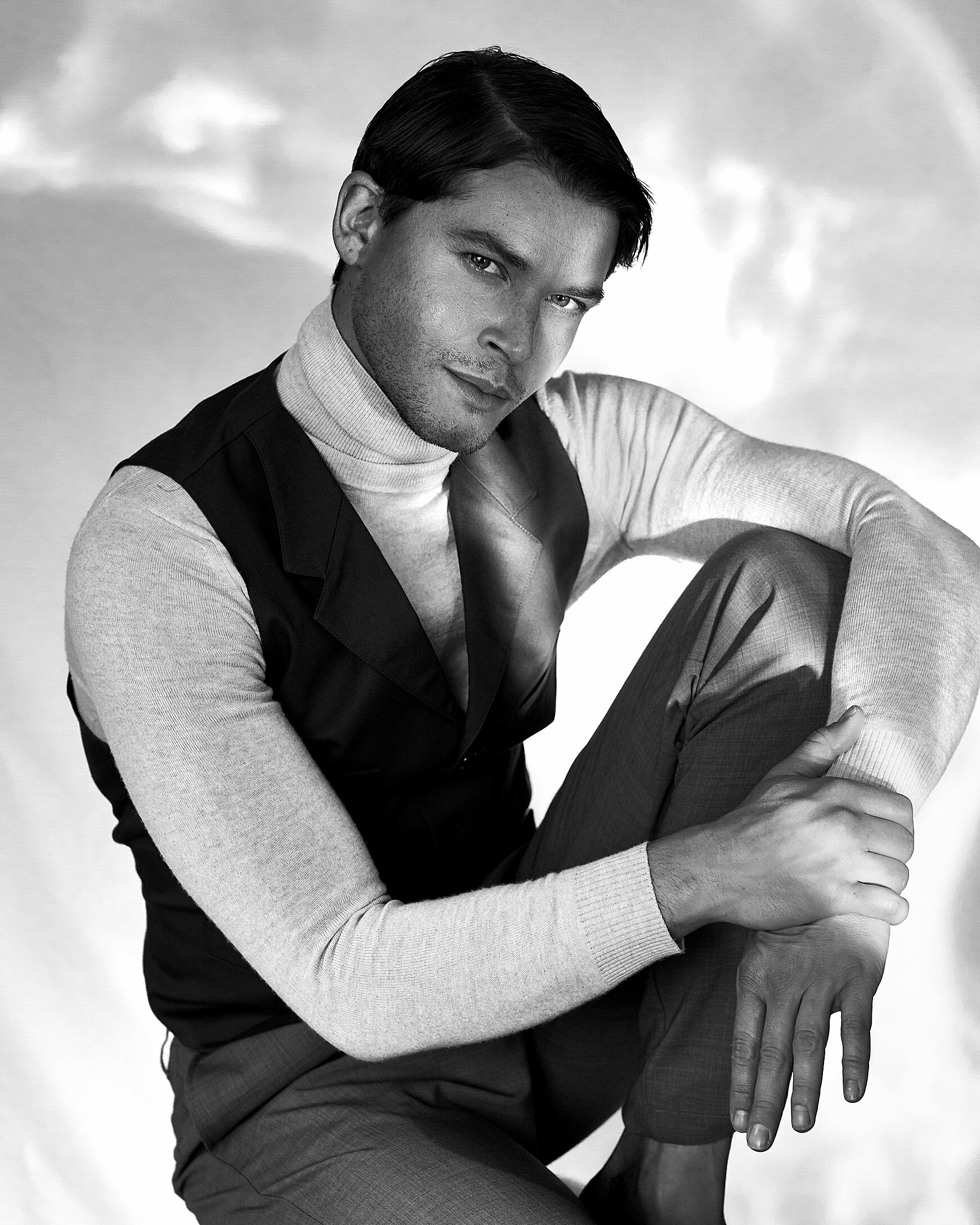 A black and white photo of a male model who wears a pullover with a turtle neck and a vest over it. He sits on a bar chair.