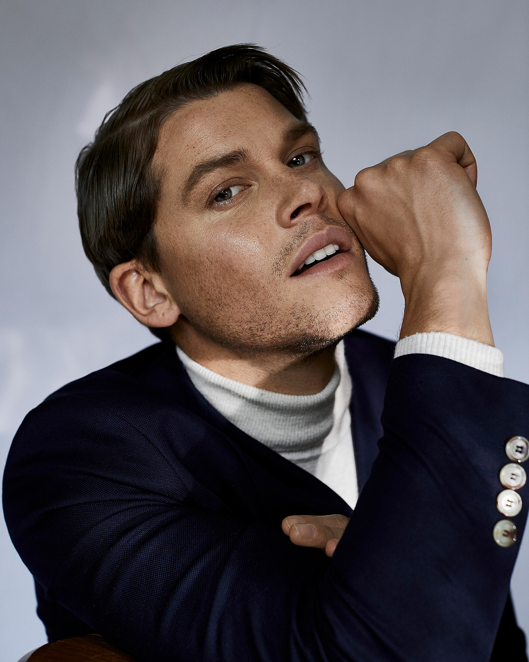 A portrait of a male model who wears a pullover with a turtle neck and a jacket over it. His hand touches his face.