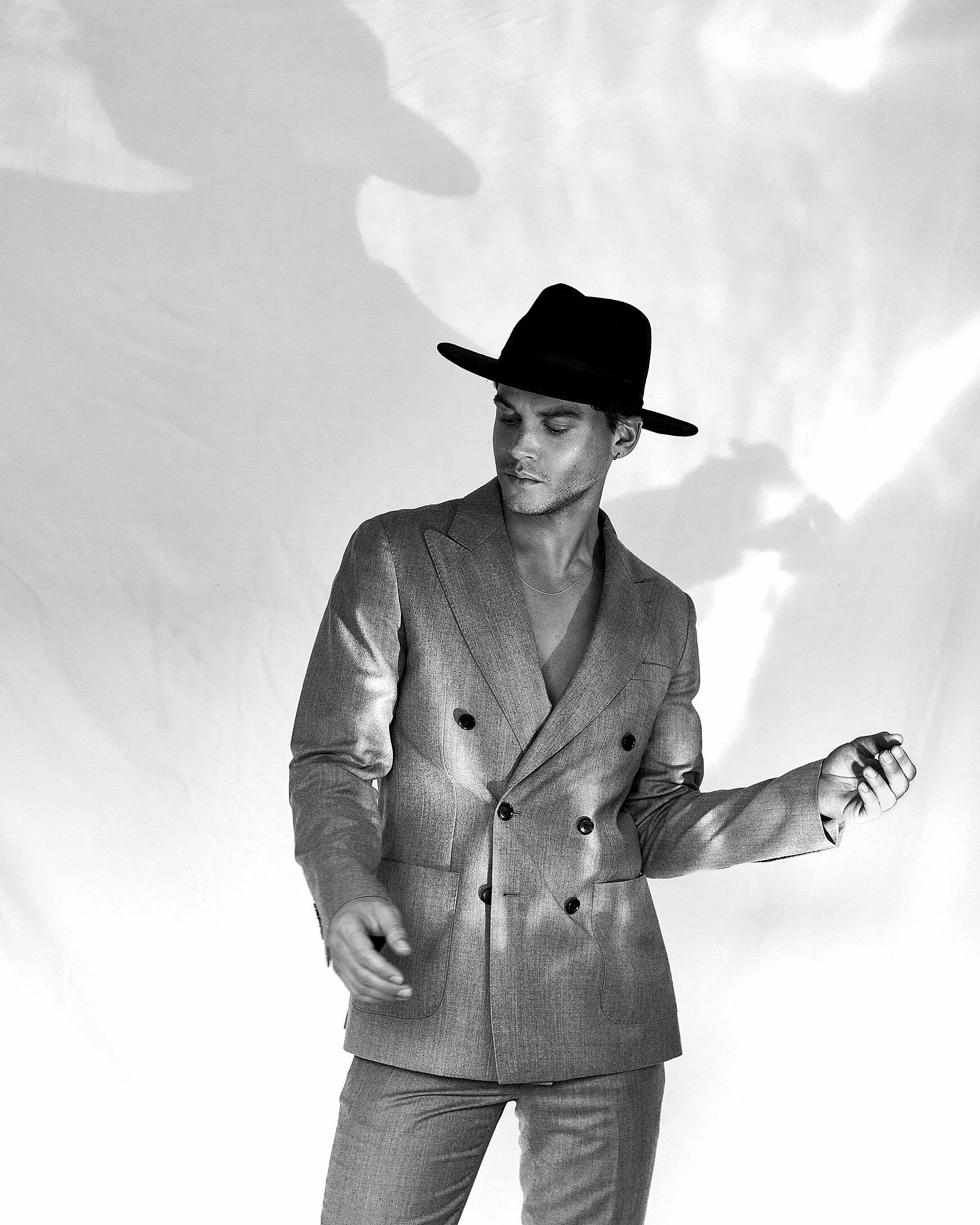 A black and white photo of a male model who wears a grey men suit and a big hat.
