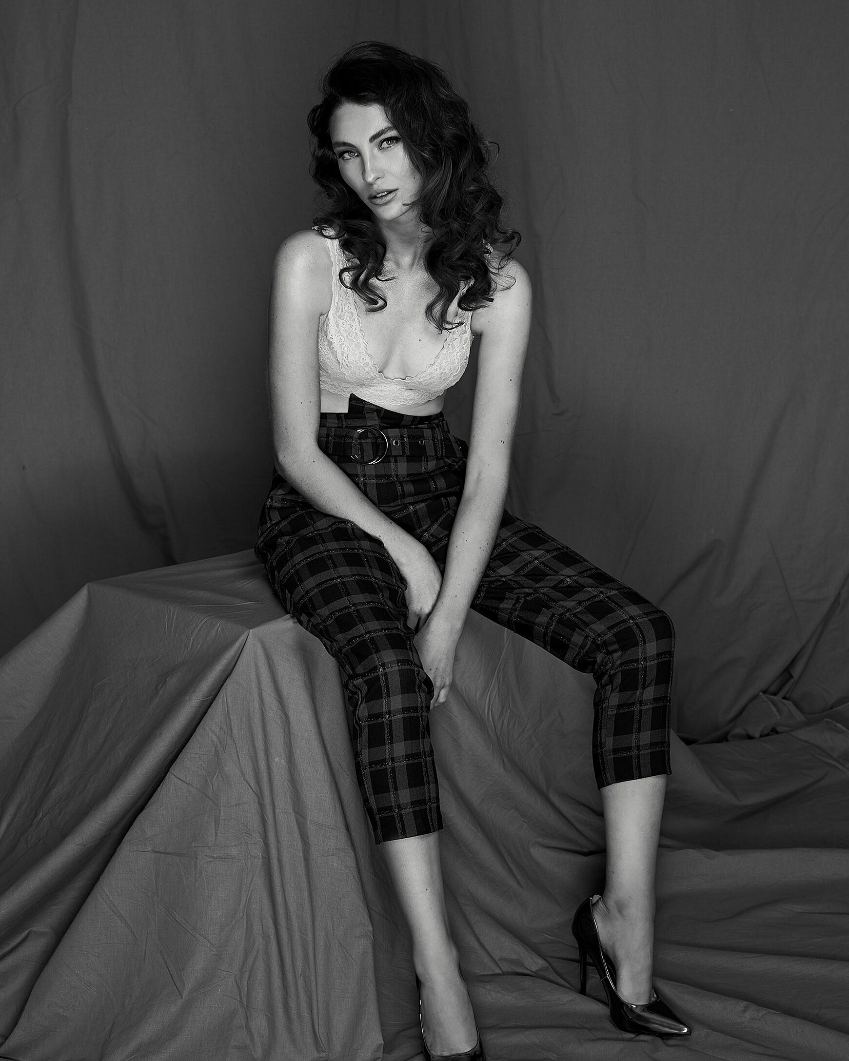 A black and white photo shows a model with brown curly hairstyle who wears a white bra and skinny knee-length trousers. 