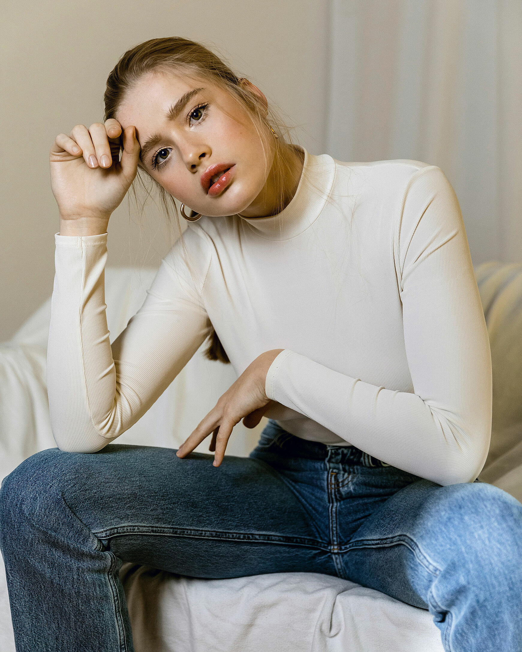 A female blonde model sits on a chair in her turtle neck white pullover and her denim trousers