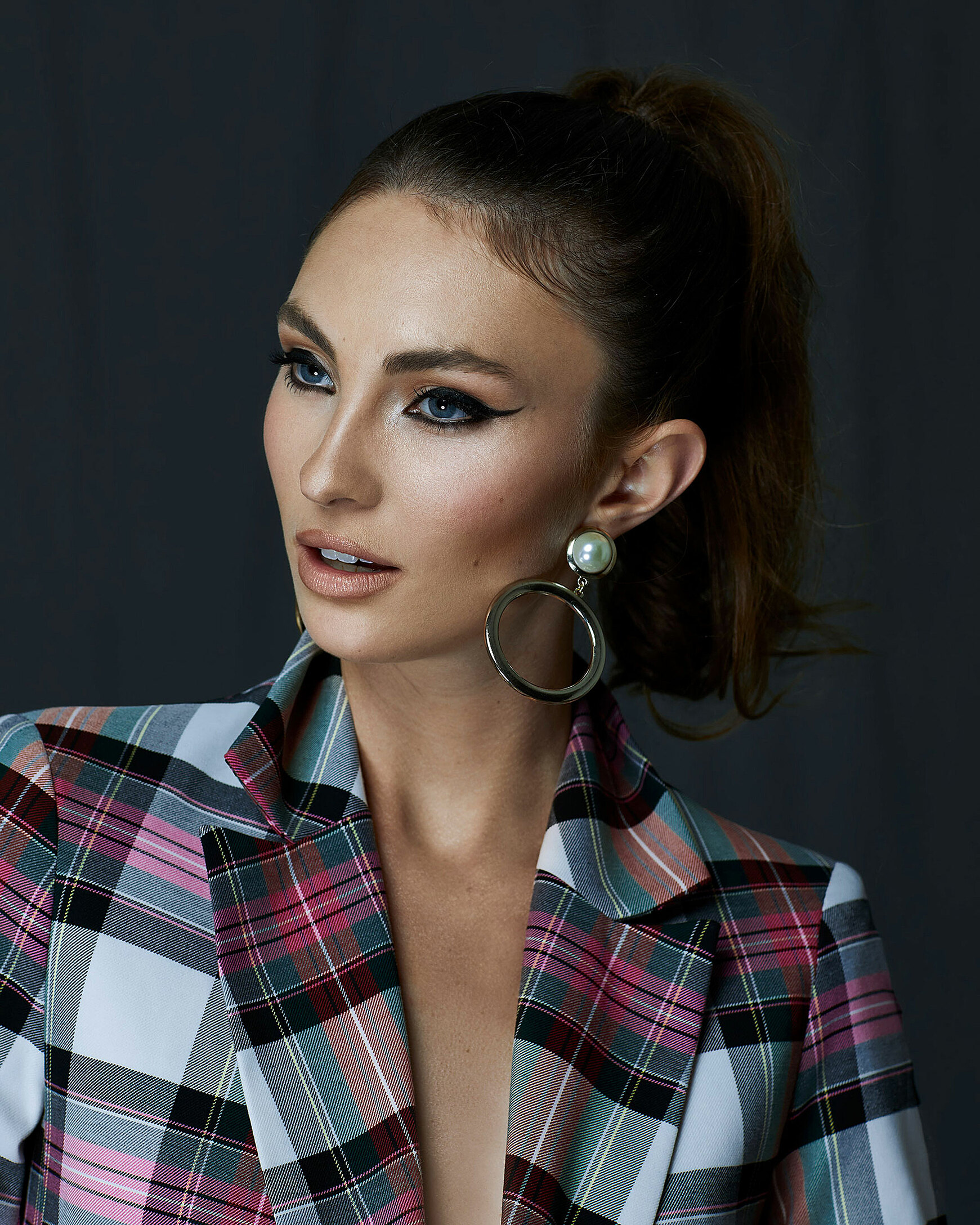 A portrait of a model with brown hair and a ponytail who wears an eyeliner and a blazer with big check pattern