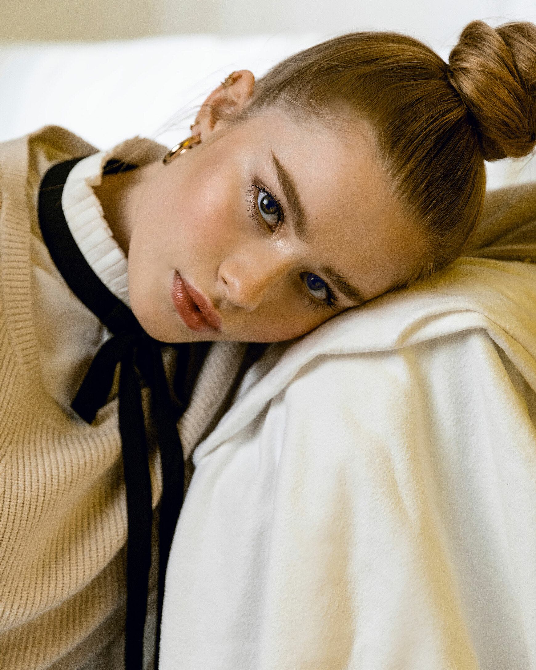 A female blonde model lean against a chair. It is a close up portrait in a beige pullover with a small black bow.