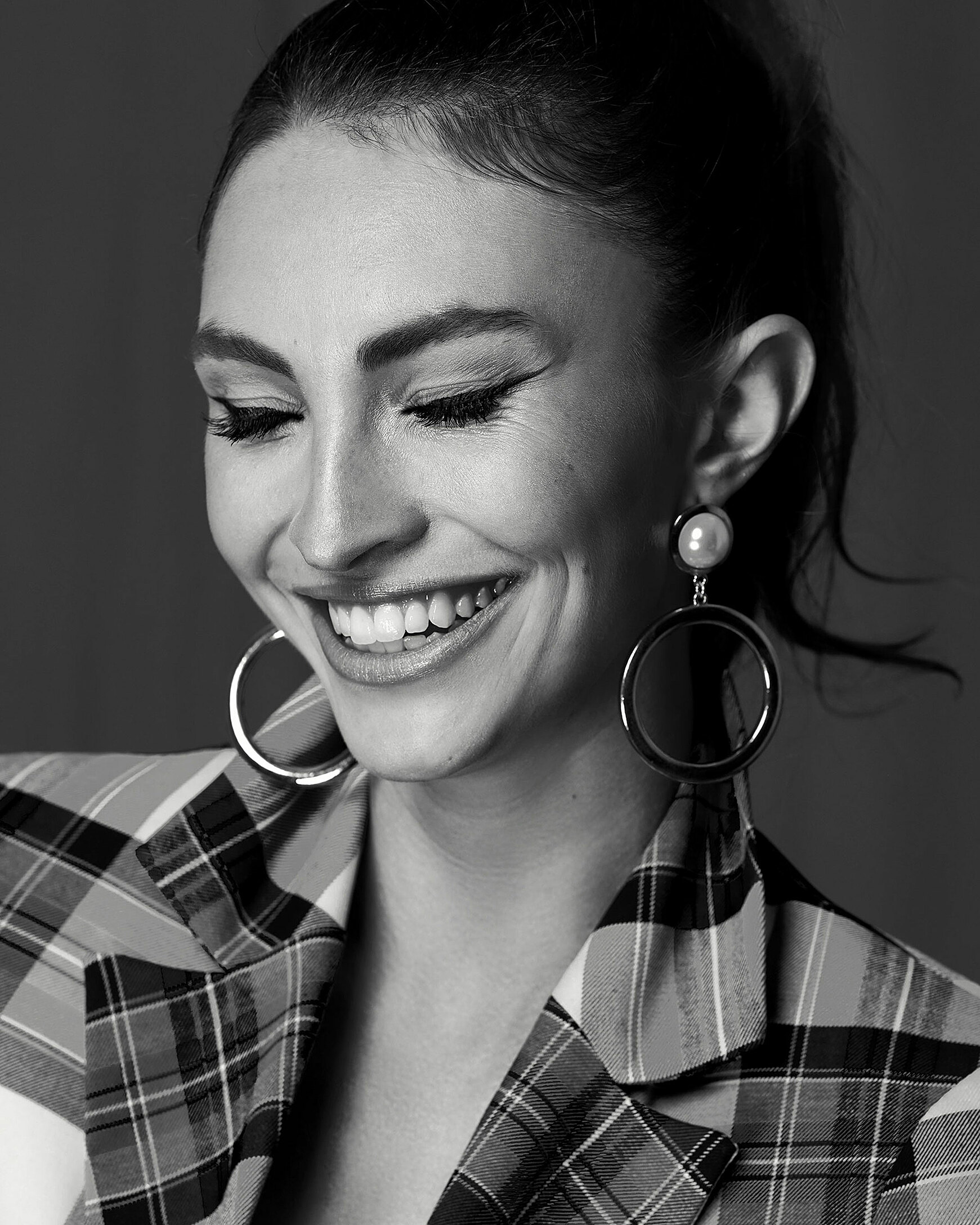 A black and white portrait of a model with brown hair and a ponytail. She wears an eyeliner and a blazer with big check pattern