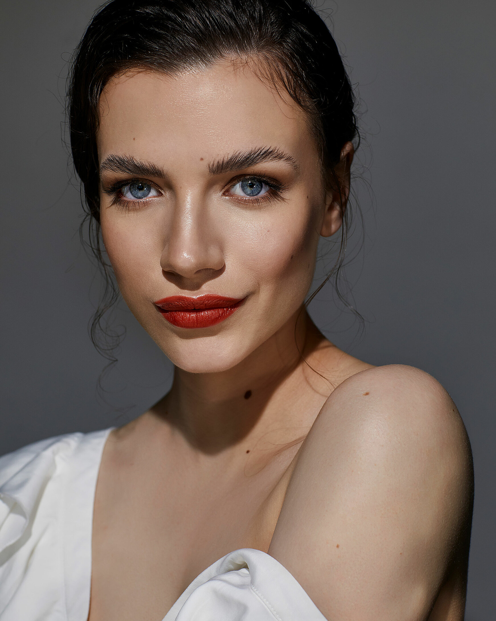 A beauty photography of a female model with dark hair. She wears a red lips and a white top