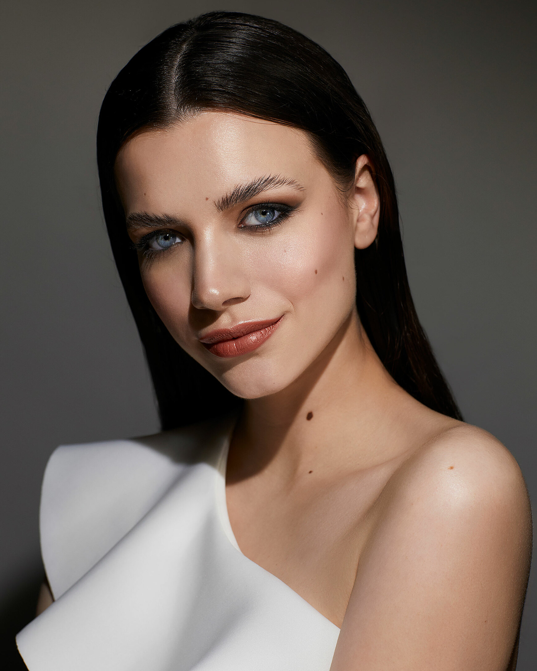 A close up of a female model with dark hair. She wears dark eyeshadow and a white dress with one shoulder.
