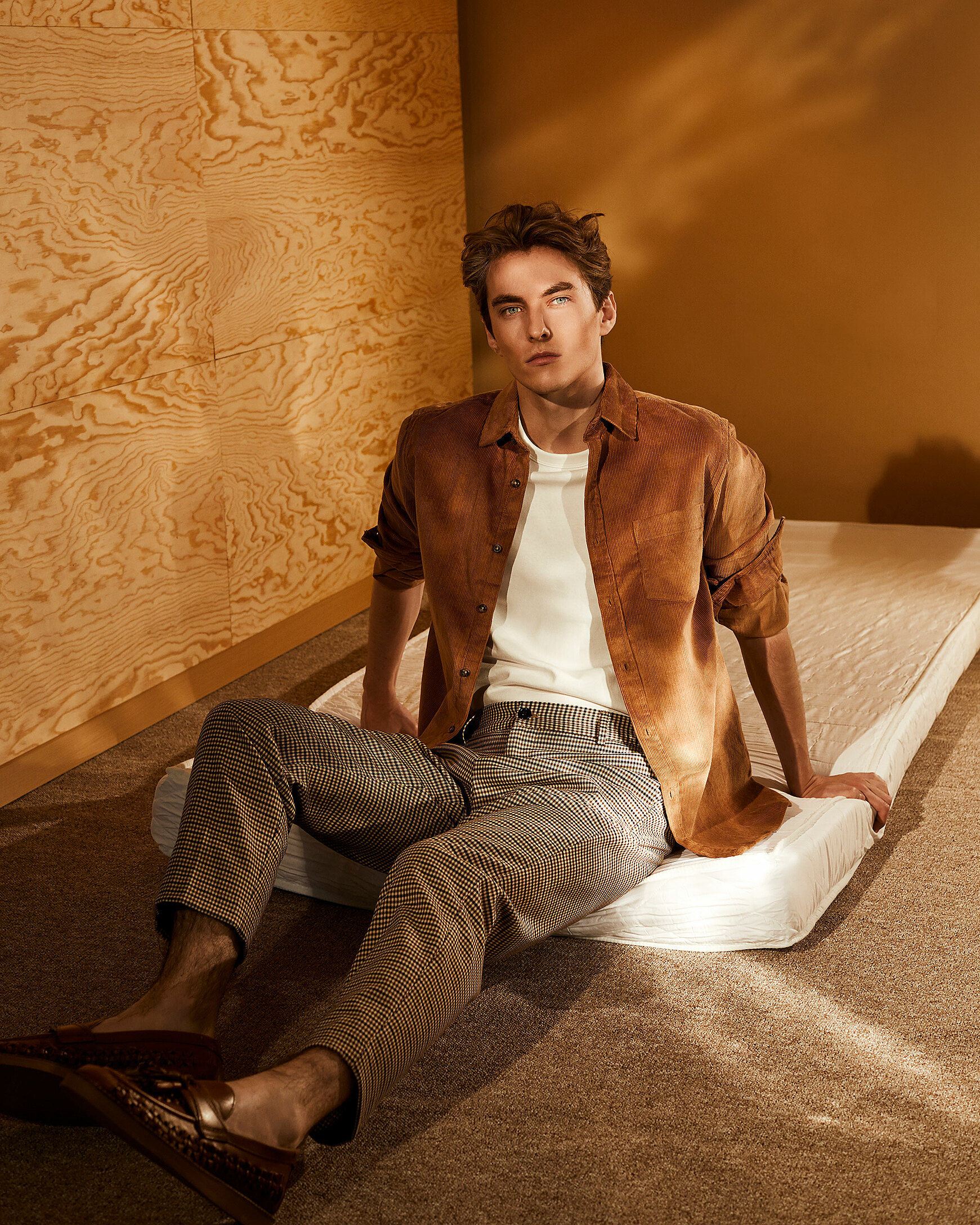 A male model sit on the mattresses in a cord jacket with a white shirt and a military green trousers.