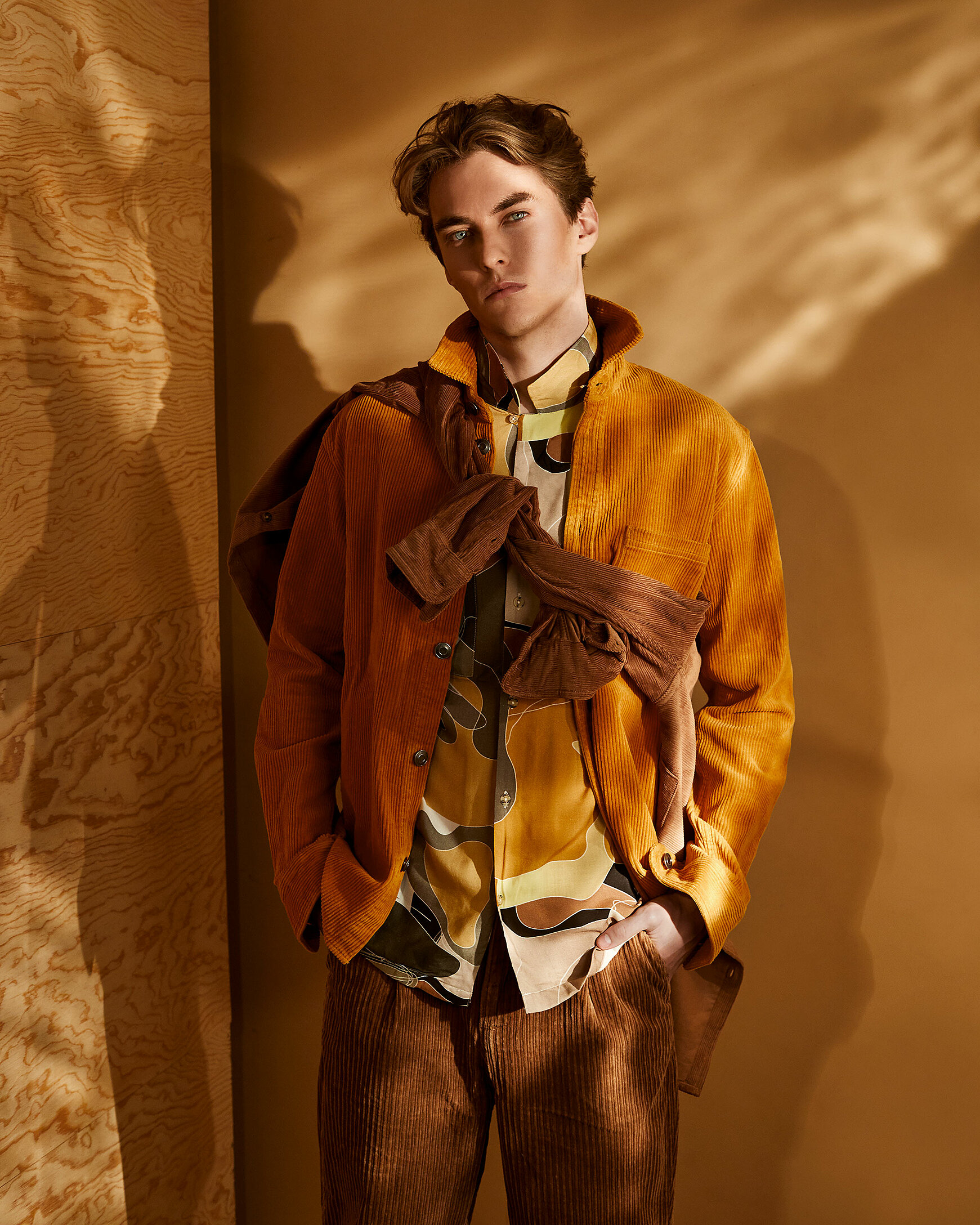 A male model with a cognac colored jacked and a patterned shirt. He wears brown cord trousers