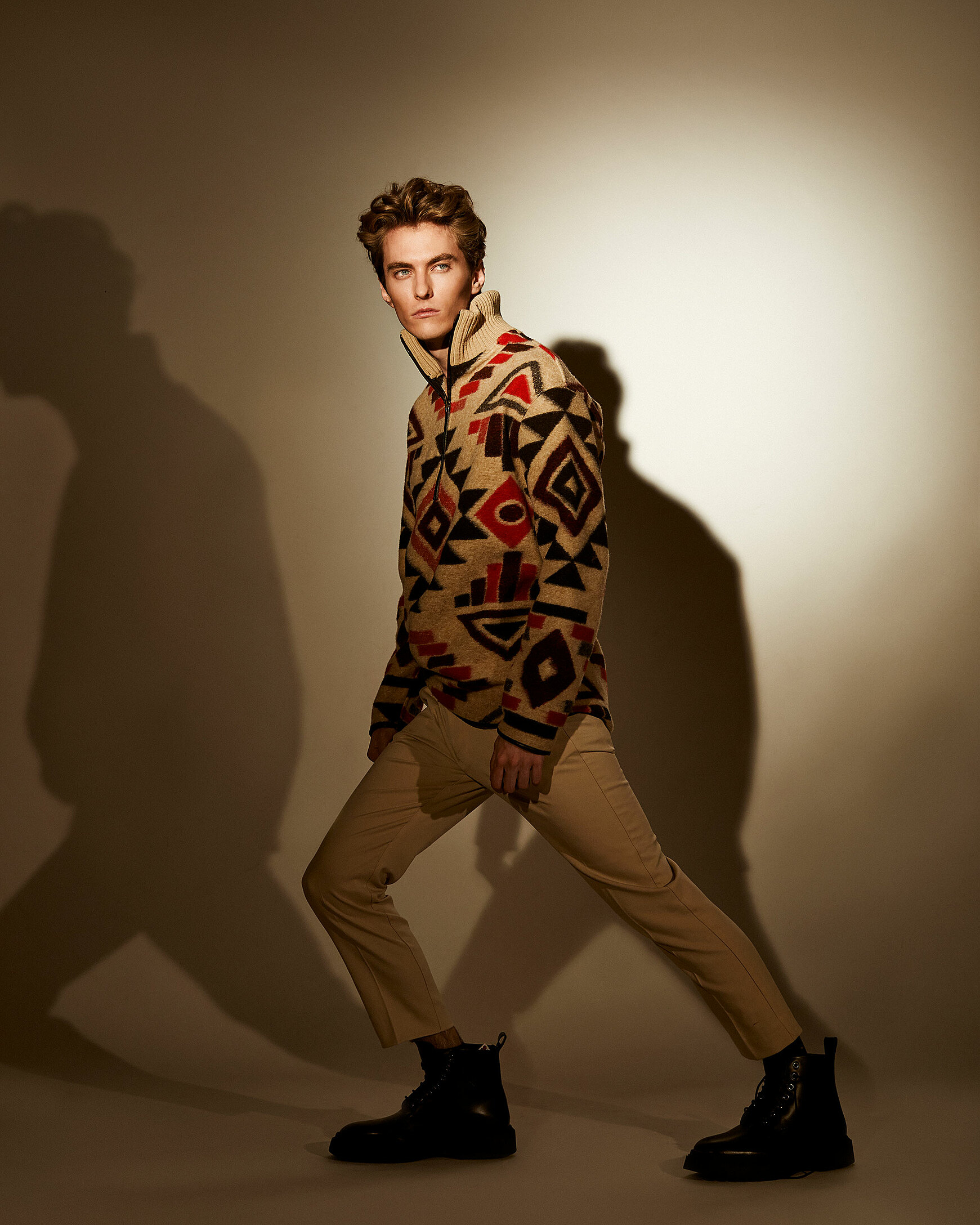 A male model with beige trousers and a patterned pullover. A hard shadow with his silhouette is in the back