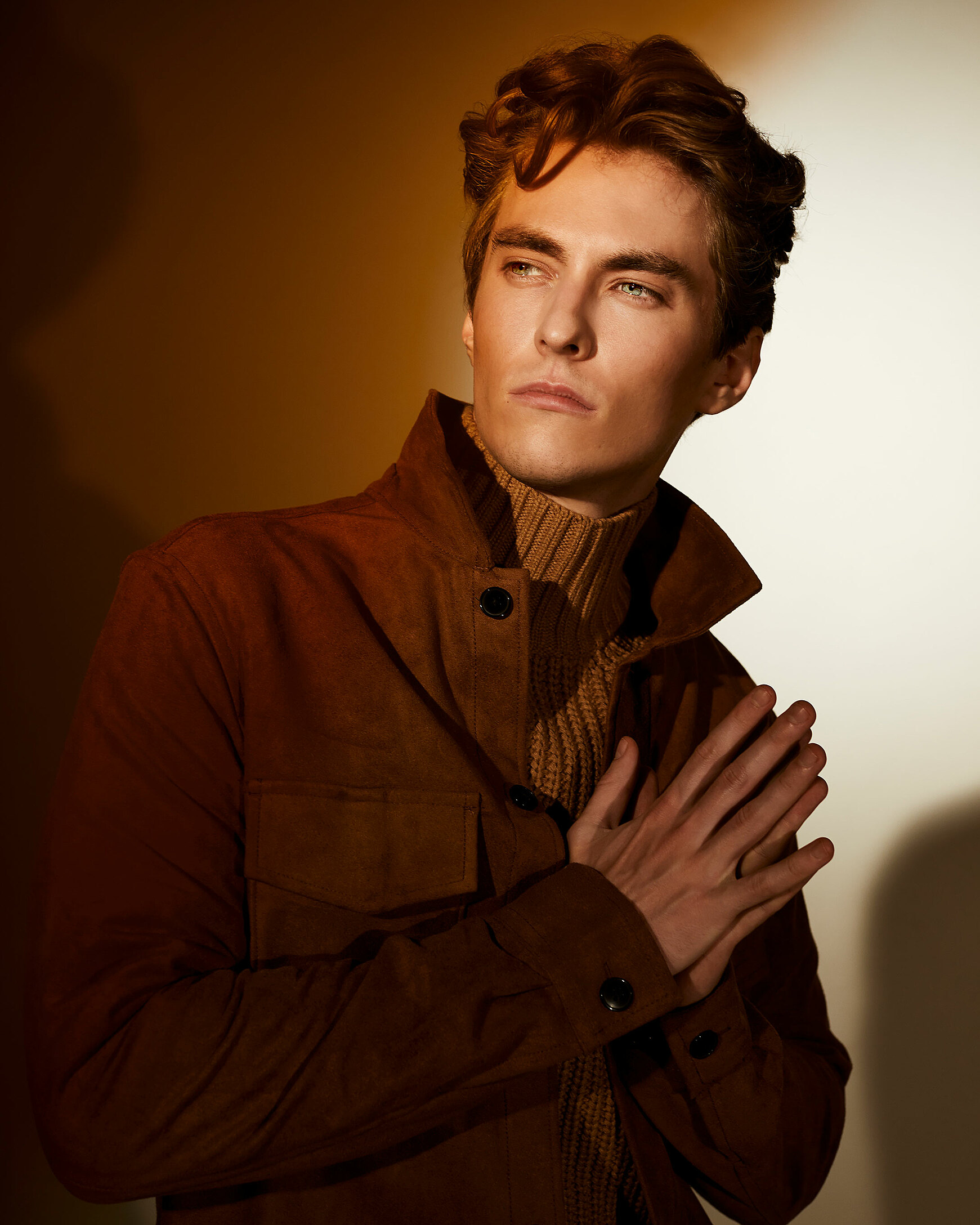 A close up of a male model who looks into the camera. It´s a dark picture with light focus on his face. He wears a pullover with a turtle neck and a brown cord jacket.