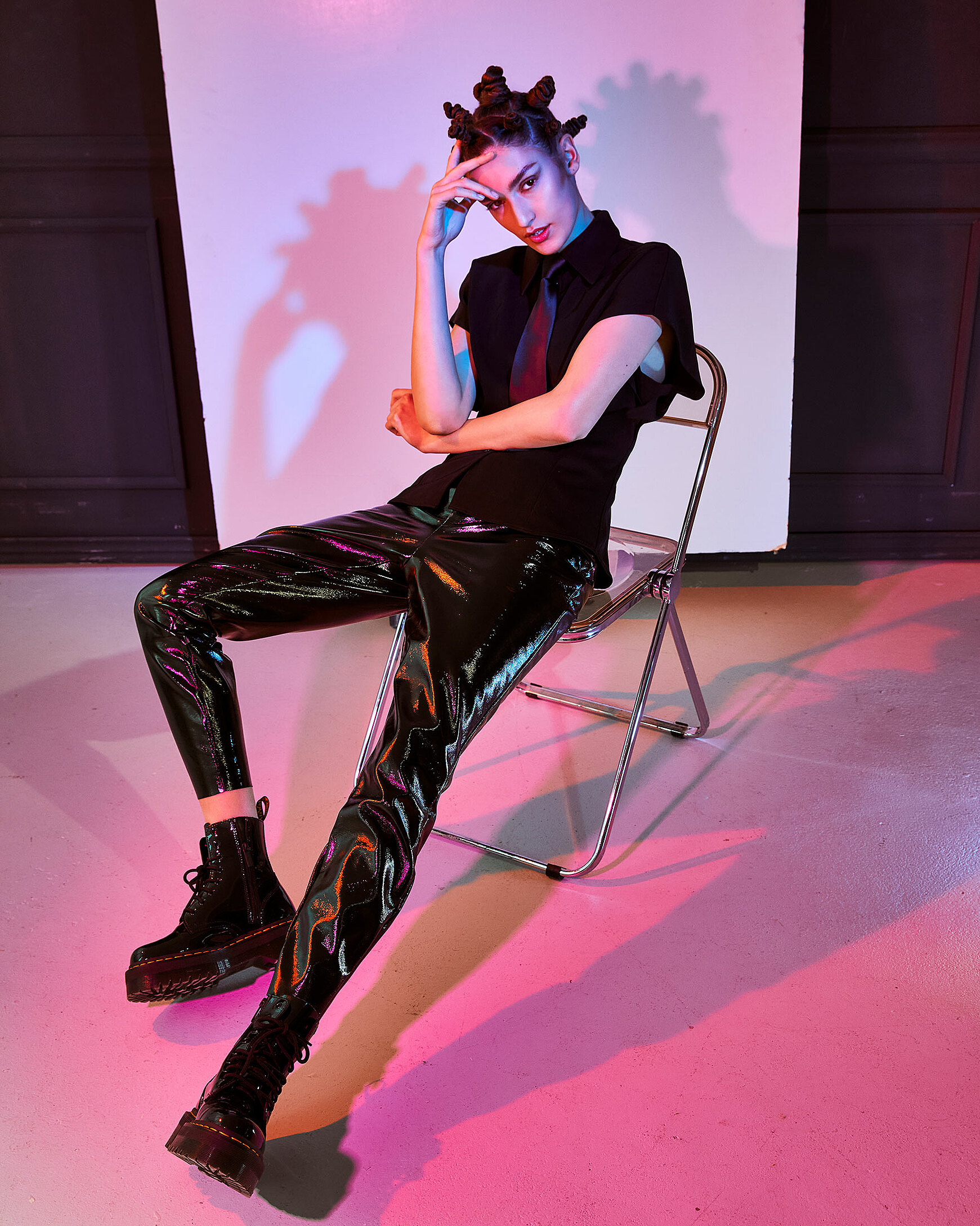 A female model sits on a transparent chair illuminated with red/ pink light in black clothes