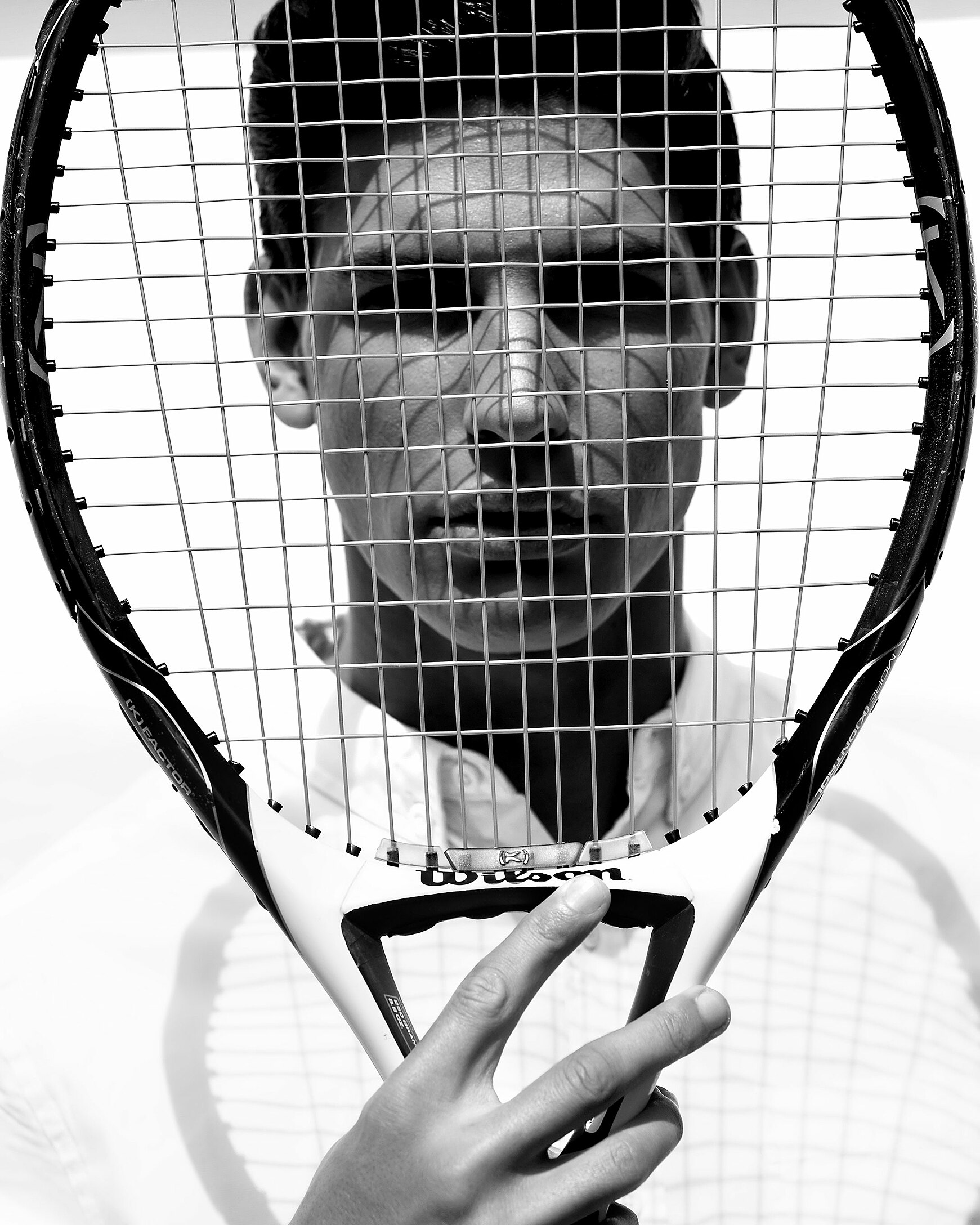 A black and white picture with a male model in a white shirt. He hold a tennis rocket in his right hand in front of his face