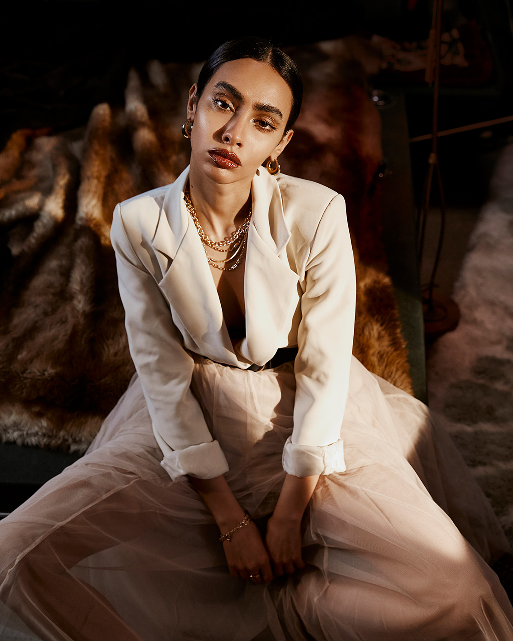 A model sits on the corner of a bed in a white blazer and rosa tull skirt
