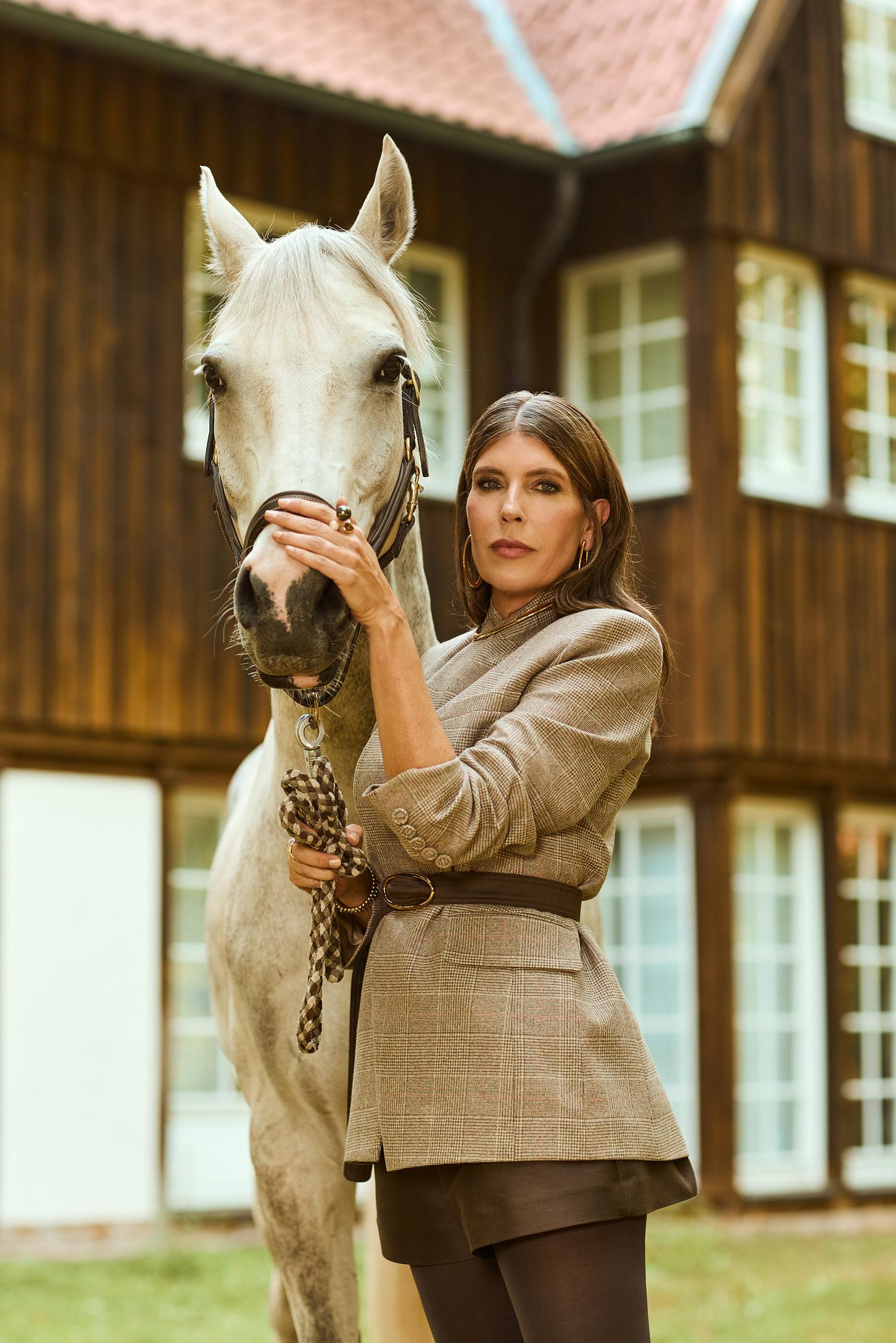 Eine Frau steht mit einem weissen Pferd in der Hand vor einem Hotel mit Holzvertäfelung und trägt einen taube farbigen Blazer
