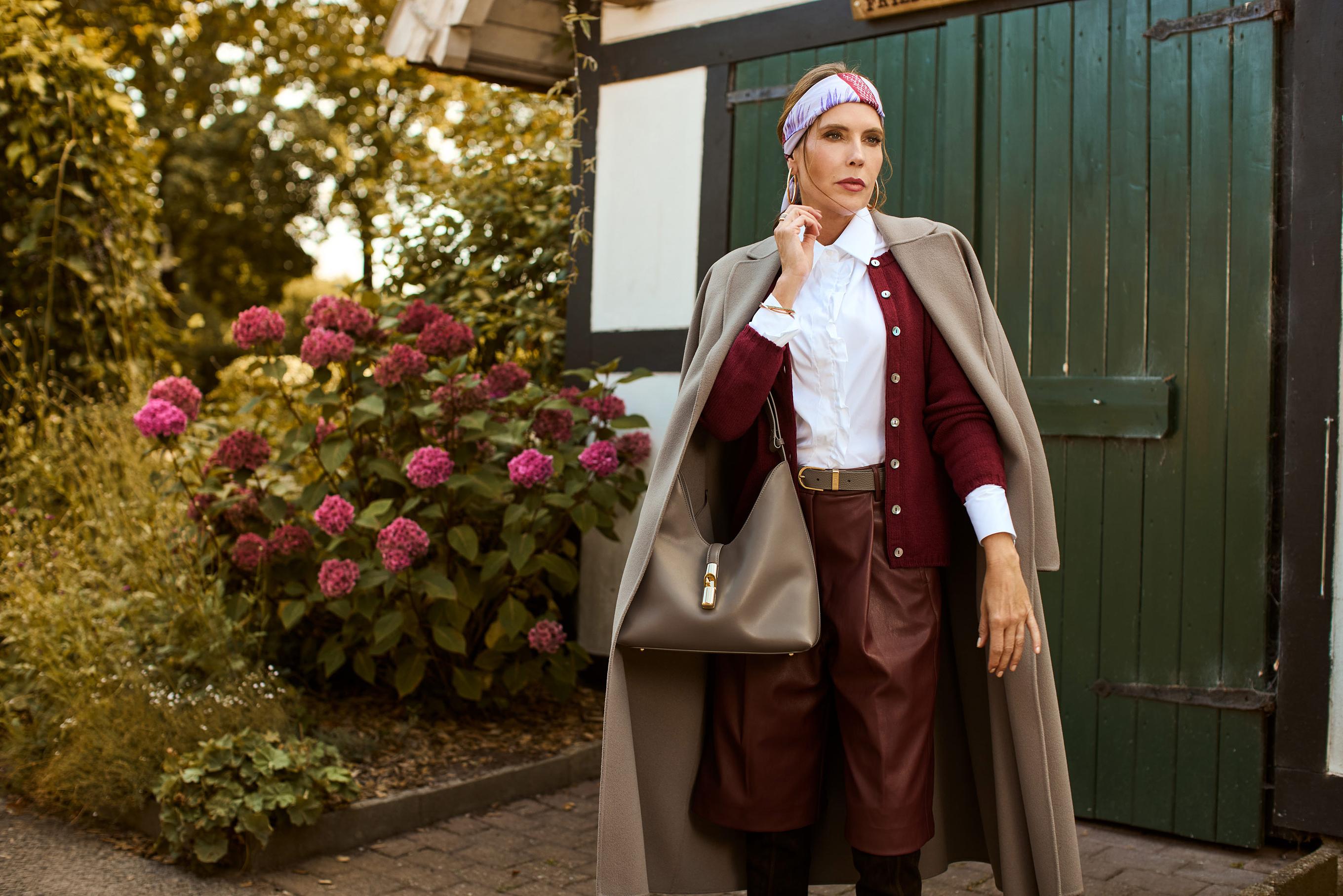 Eine Frau trägt eine Kombination aus einer dunkel roten Culotte und einer ebenso dunkel roten Strickjacke. Darüber trägt Sie einen Mantel in taupe und hält eine Tasche. Ihr Styling ist mit einem violetten Stirnband abgerundet.