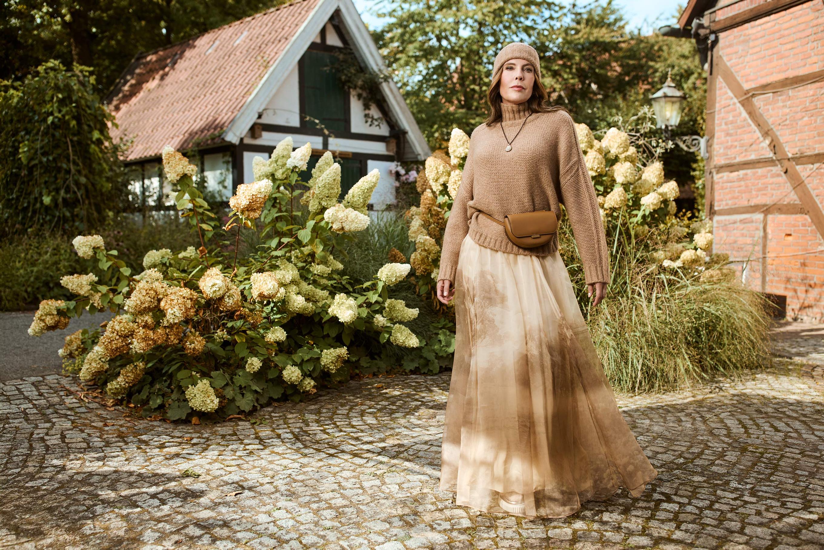 Eine Umgebung zwischen Hortensien und Fachwerkhäusern. Dazwischen steht eine Frau in einem einem zarten hell braunen Rock mit einer Wollmütze und einem hell braunen Pullover 