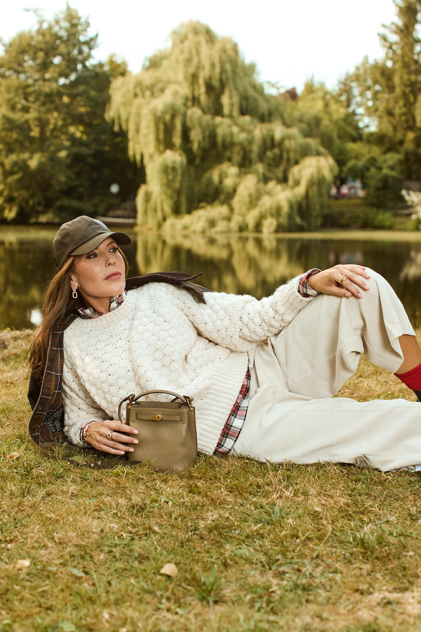 Eine Frau liegt auf einer Wiese vor einem See, Natur ist im Hintergrund zu sehen und sie posiert in beiger Mode mit einer Tasche.