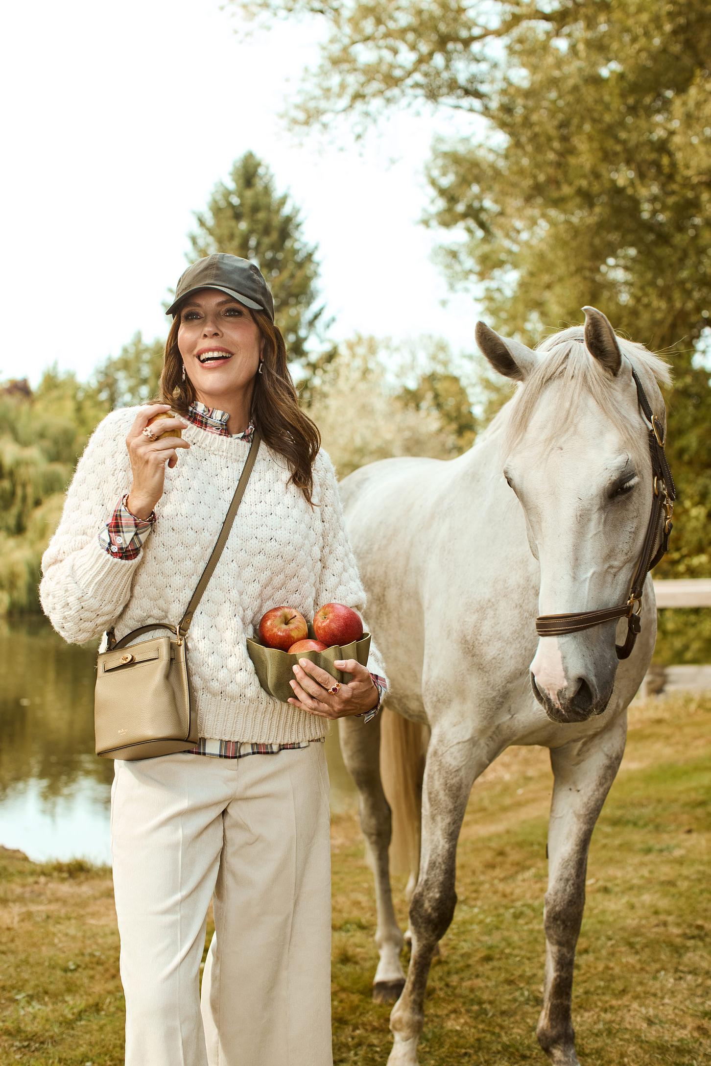 Eine Frau steht mit einer Schale Äpfel vor einem weissen Pferd und lächelt. Natur ist im Hintergrund zu sehen und sie posiert in beiger Mode mit einer Tasche.