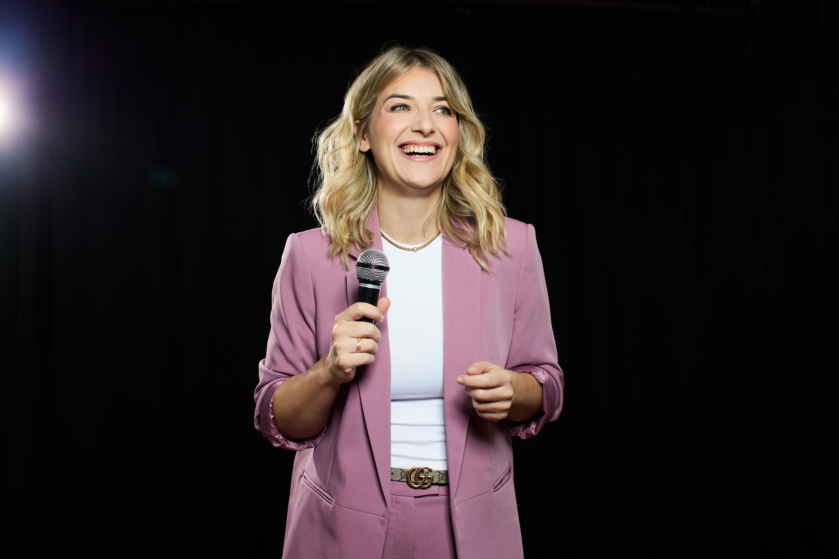 Frau im rosé farbigen Blazer steht mit Mikrofon auf einer Bühne. Gegenlicht im Hintergrund