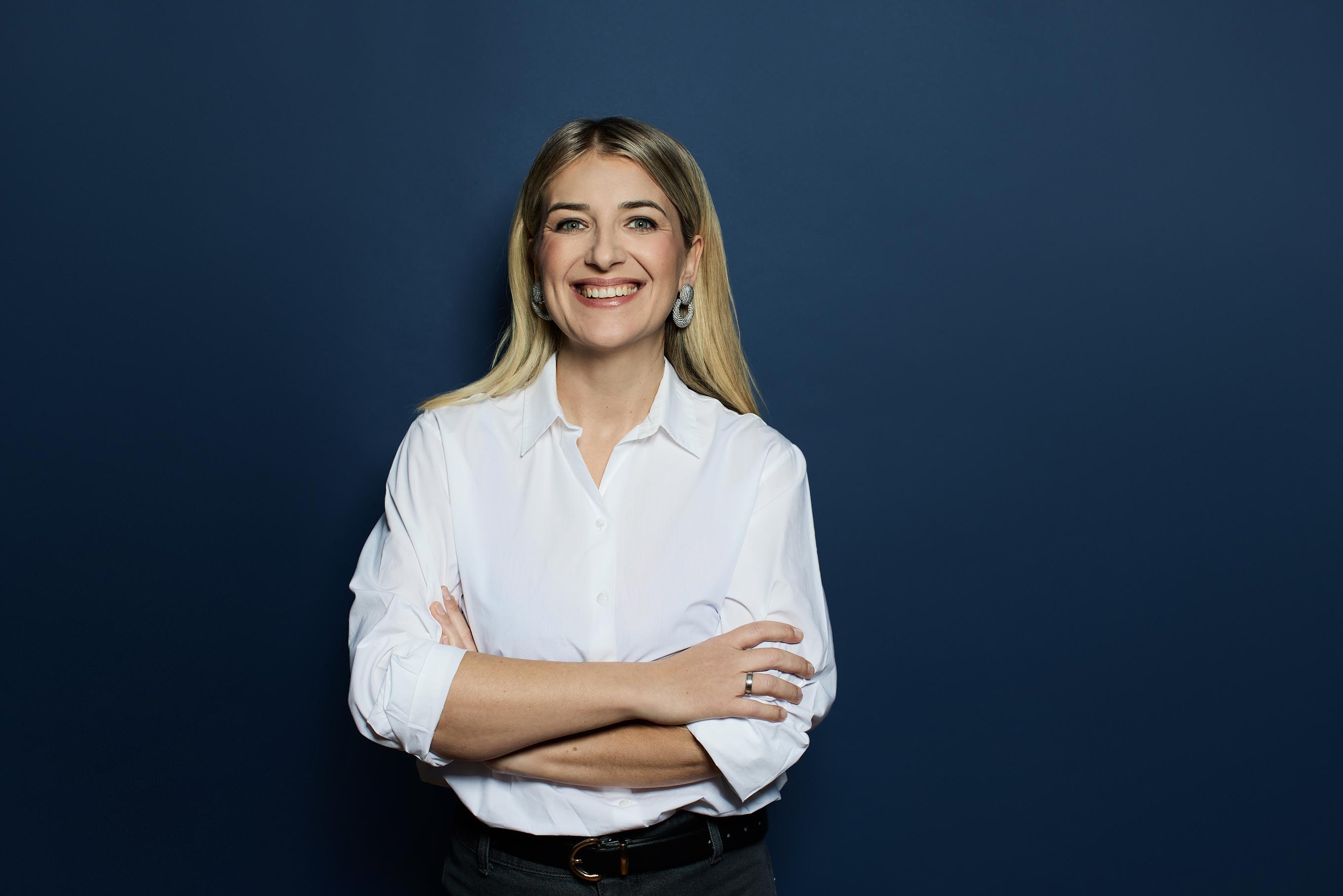 Frau in einer weissen Bluse lacht und steht angelehnt gegen einer blauen Wand