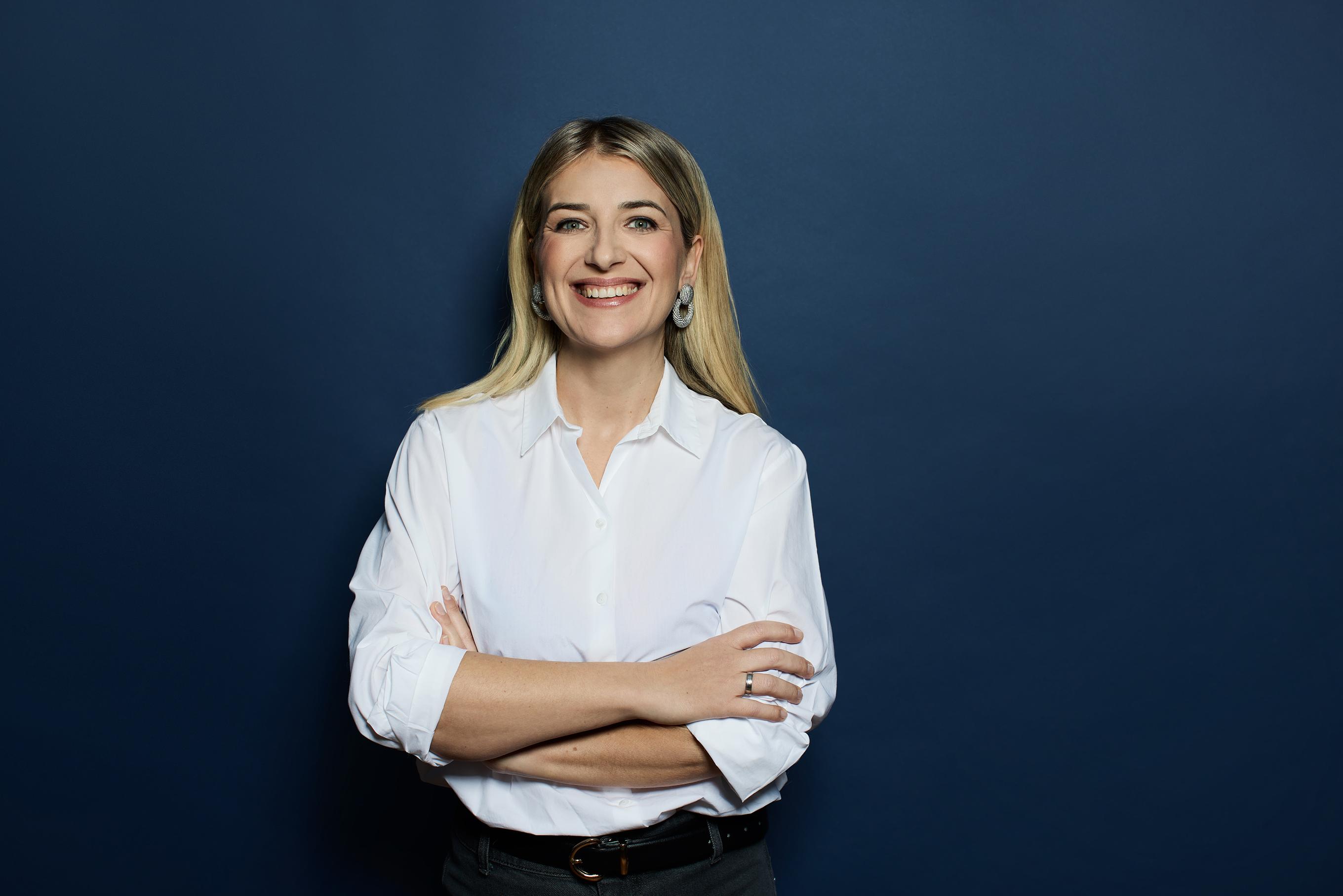 Frau in einer weissen Bluse lacht und steht angelehnt gegen einer blauen Wand