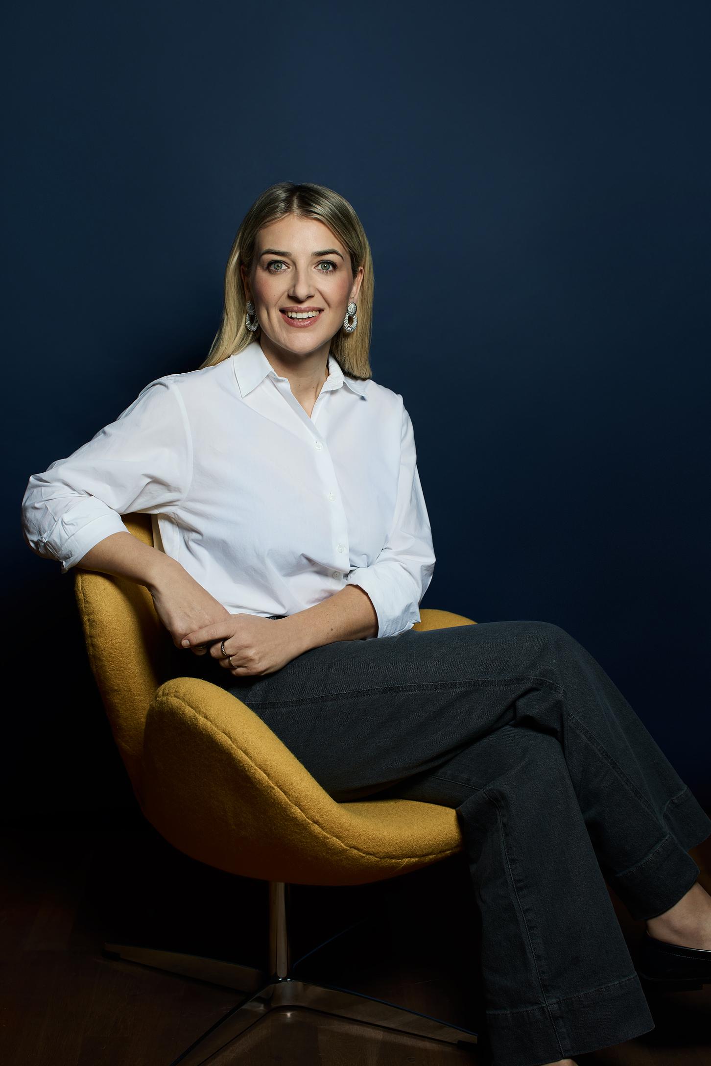 Frau in einer weissen Bluse sitzt auf einem gelben Stuhl vor einer blauen Wand