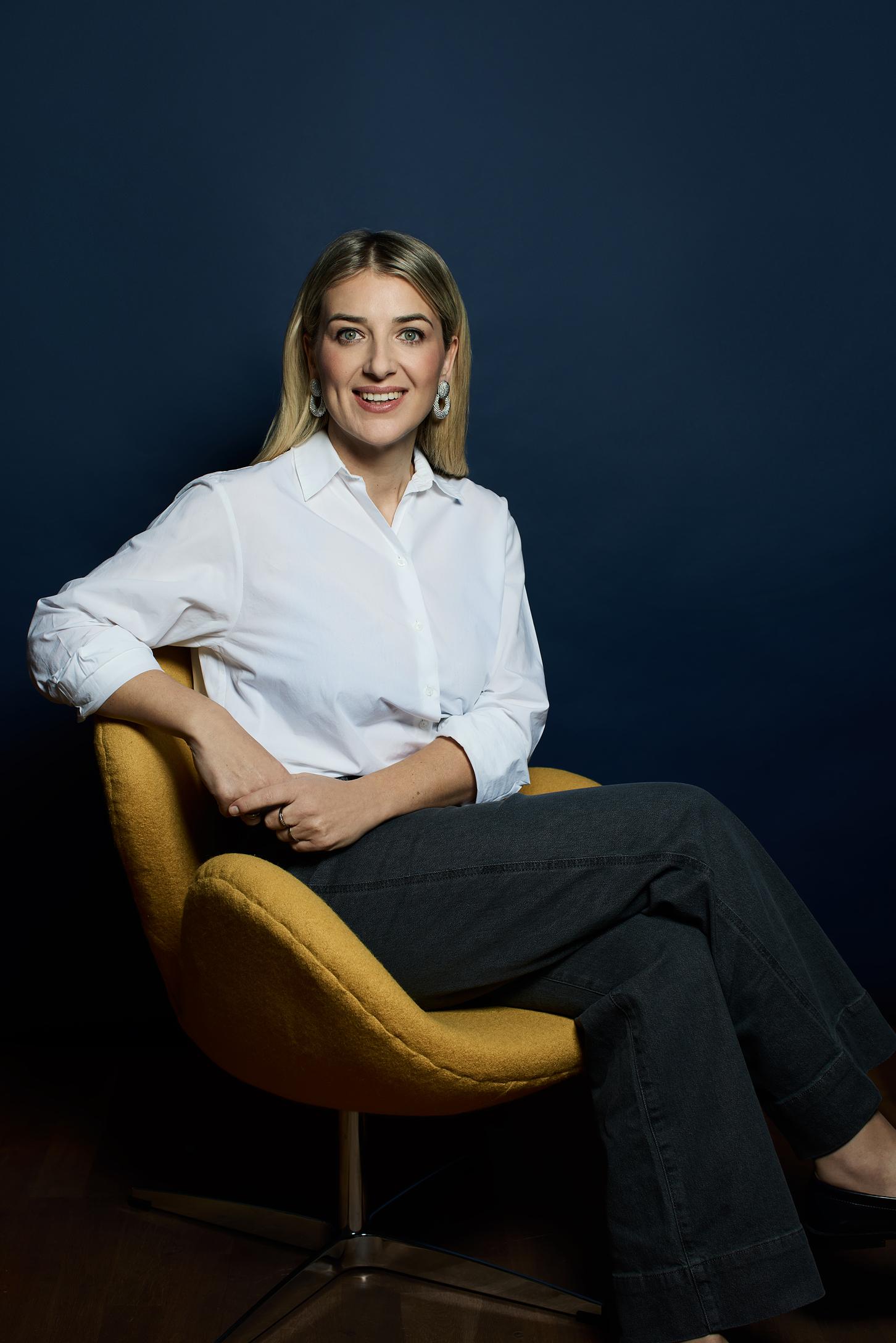 Frau in einer weissen Bluse sitzt auf einem gelben Stuhl vor einer blauen Wand