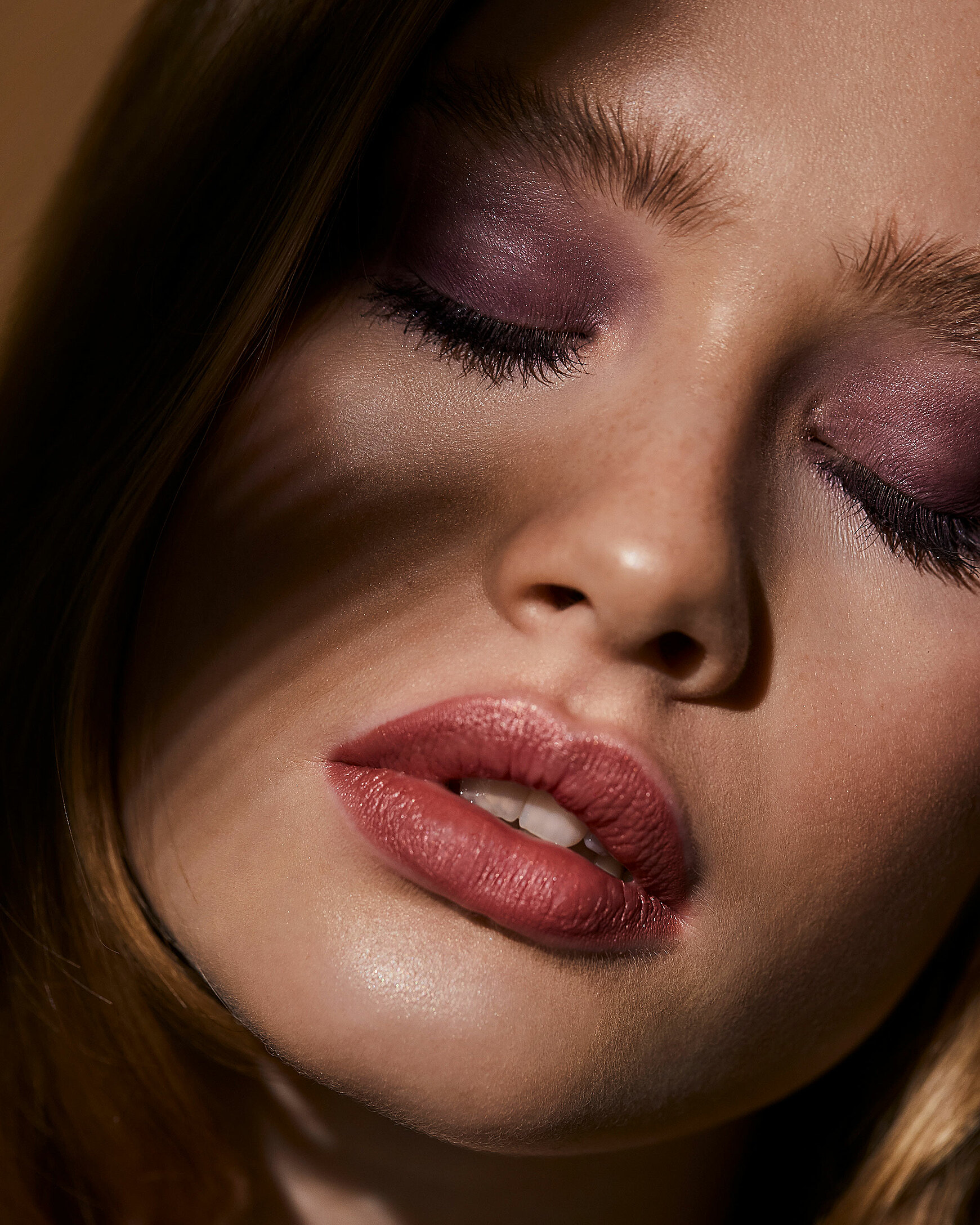 A close-up of a blonde model with delicate red lips and purple eye shadow